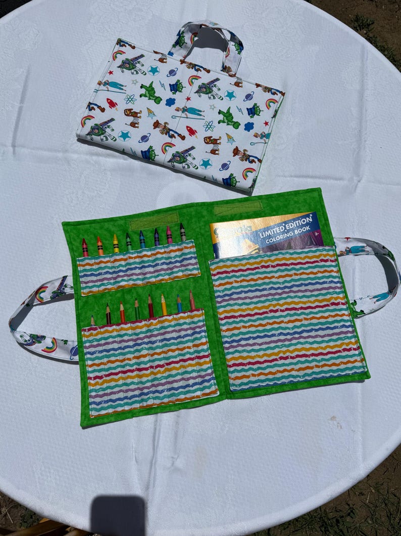 May include: A fabric carrying case with a white exterior featuring a colorful cartoon character print and a bright green interior. The case unfolds to reveal multiple slots holding colored pencils and a pocket containing a limited edition coloring book.