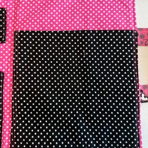 Pu&ograve; includere: Un organizer in tessuto con sfondo rosa acceso a pois bianchi e una grande tasca centrale nera a pois bianchi. L'articolo include una striscia di chiusura a strappo nera rettangolare sulla parte superiore e linguette laterali con stampe di personaggi dei cartoni.