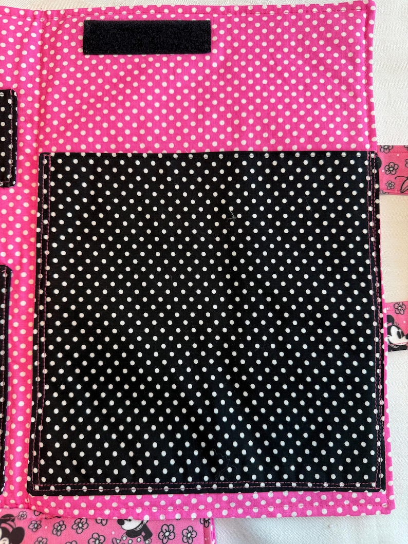 Pu&ograve; includere: Un organizer in tessuto con sfondo rosa acceso a pois bianchi e una grande tasca centrale nera a pois bianchi. L'articolo include una striscia di chiusura a strappo nera rettangolare sulla parte superiore e linguette laterali con stampe di personaggi dei cartoni.