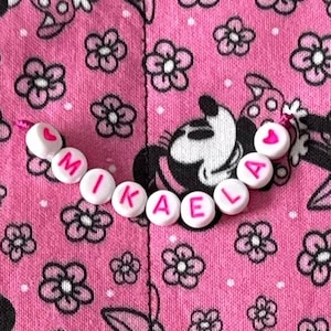 May include: A string of white plastic beads with pink lettering spelling the name MIKAELA, flanked by two small pink heart beads. The beads are arranged in a curved line against a pink fabric background featuring a black floral and character pattern.