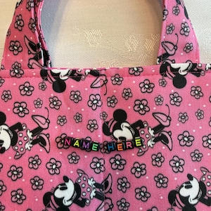 Pu&ograve; includere: Una borsa in tessuto rosa con un motivo ripetuto di personaggi di topo dei cartoni animati in bianco e nero e piccoli fiori. La borsa include due manici in tessuto e una fila di perline con lettere in plastica colorata che compongono il testo NAME HERE.