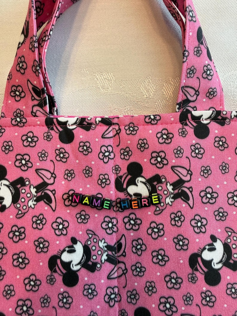 Pu&ograve; includere: Una borsa in tessuto rosa con un motivo ripetuto di personaggi di topo dei cartoni animati in bianco e nero e piccoli fiori. La borsa include due manici in tessuto e una fila di perline con lettere in plastica colorata che compongono il testo NAME HERE.