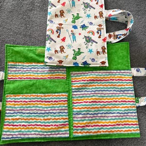 May include: A fabric organizer case featuring a white exterior with a colorful Toy Story character print and a bright green interior. The interior includes multiple pockets with a wavy, multi-colored striped pattern and a fabric handle.