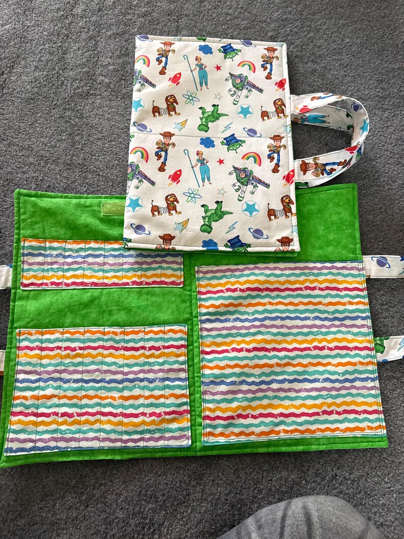 May include: A fabric organizer case featuring a white exterior with a colorful Toy Story character print and a bright green interior. The interior includes multiple pockets with a wavy, multi-colored striped pattern and a fabric handle.