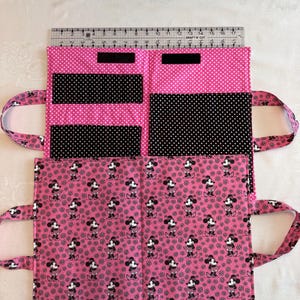 Pu&ograve; includere: Un organizer in tessuto rosa e nero con motivo Minnie Mouse, dotato di tasche interne e due manici. Un righello in plastica trasparente &egrave; posizionato lungo il bordo superiore, mostrando una larghezza di circa 43 cm per l'organizer aperto.