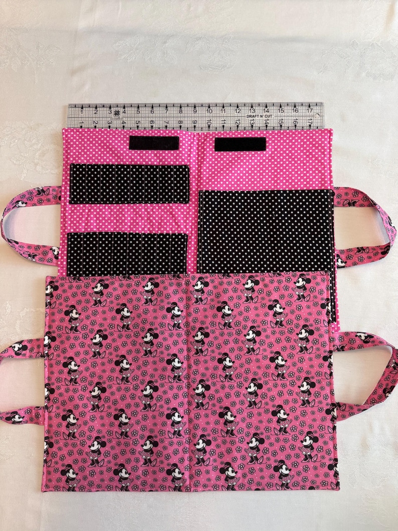 Pu&ograve; includere: Un organizer in tessuto rosa e nero con motivo Minnie Mouse, dotato di tasche interne e due manici. Un righello in plastica trasparente &egrave; posizionato lungo il bordo superiore, mostrando una larghezza di circa 43 cm per l'organizer aperto.