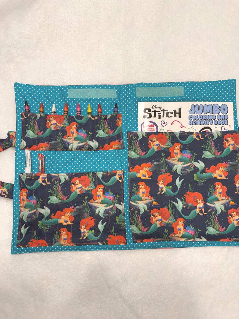 May include: A fabric activity organizer featuring a teal background with white polka dots and a dark blue pattern of Ariel the mermaid. It includes pockets holding crayons and markers, and a Disney Stitch Jumbo Coloring and Activity Book.