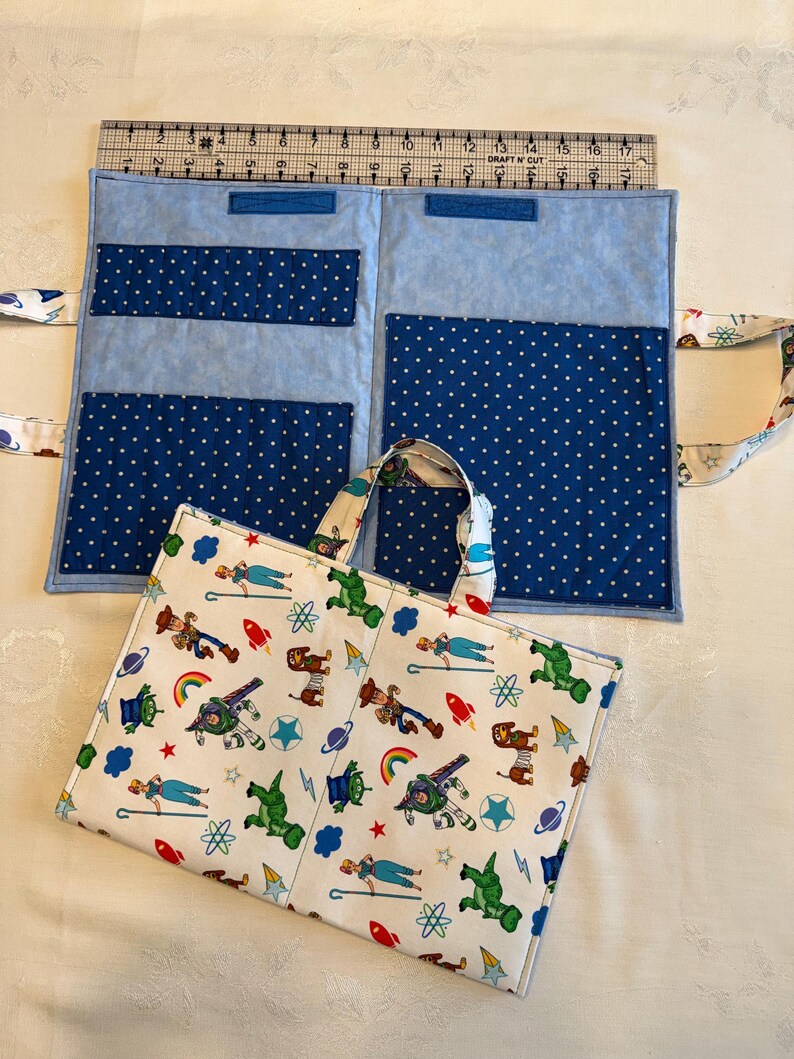 May include: A fabric organizer with a white exterior featuring a colorful cartoon character print and a light blue interior with dark blue polka-dot pockets. A metal ruler placed along the top edge shows the item measures 17 inches in width.