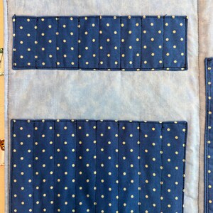 May include: A light blue fabric organizer featuring a mottled pattern with two dark blue rectangular pockets adorned with white polka dots. The pockets are stitched into vertical slots for storage, and a dark blue velcro strap is at the top.