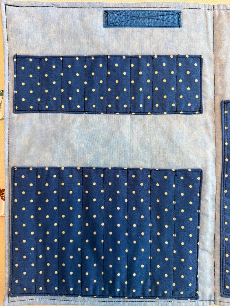 May include: A light blue fabric organizer featuring a mottled pattern with two dark blue rectangular pockets adorned with white polka dots. The pockets are stitched into vertical slots for storage, and a dark blue velcro strap is at the top.