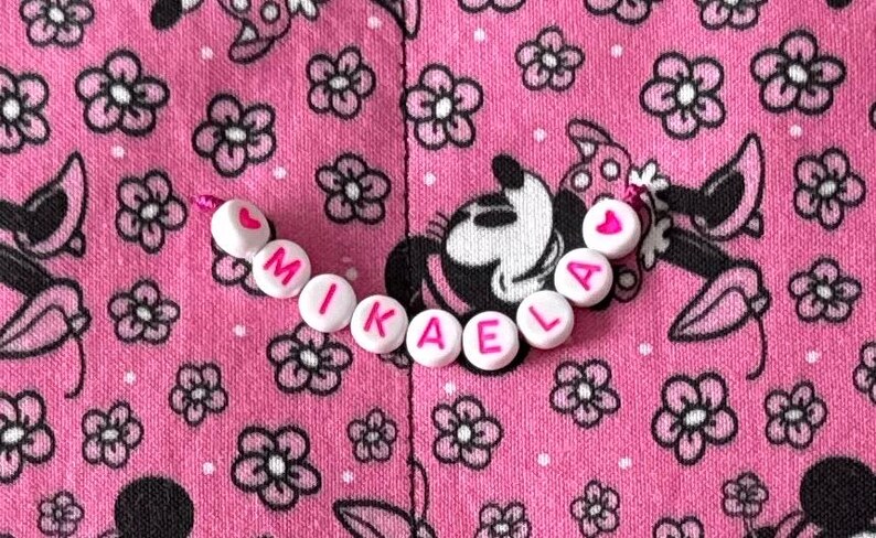 Pu&ograve; includere: Una fila di perline di plastica bianca con lettere rosa che compongono il nome MIKAELA, affiancata da due piccole perline a forma di cuore rosa. Le perline sono disposte in linea curva su un tessuto rosa con un motivo floreale e di personaggi.