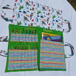 May include: A fabric coloring book organizer featuring a white exterior with a Toy Story character print and a green interior. The interior includes pockets holding colored pencils and a Crayola Limited Edition Coloring Book with a yellow cover.