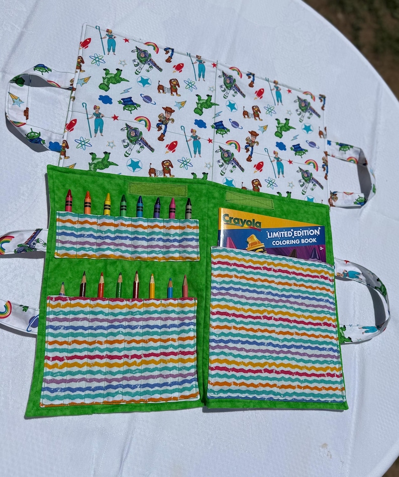 May include: A fabric coloring book organizer featuring a white exterior with a Toy Story character print and a green interior. The interior includes pockets holding colored pencils and a Crayola Limited Edition Coloring Book with a yellow cover.