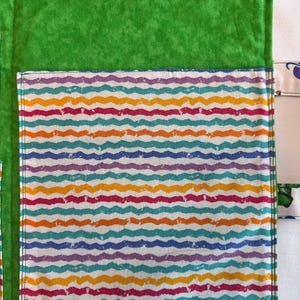May include: A bright green fabric organizer featuring a large rectangular pocket with a repeating pattern of horizontal wavy lines in shades of purple, orange, teal, and pink. The organizer includes a green hook-and-loop fastener strip at the top.
