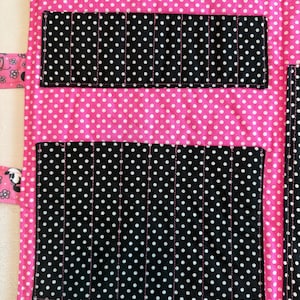 Pu&ograve; includere: Un organizer in tessuto con una base rosa brillante a pois bianchi. Include diverse tasche nere a pois bianchi per riporre oggetti, una striscia di velcro nera nella parte superiore e un bordo rosa con un motivo floreale in bianco e nero.