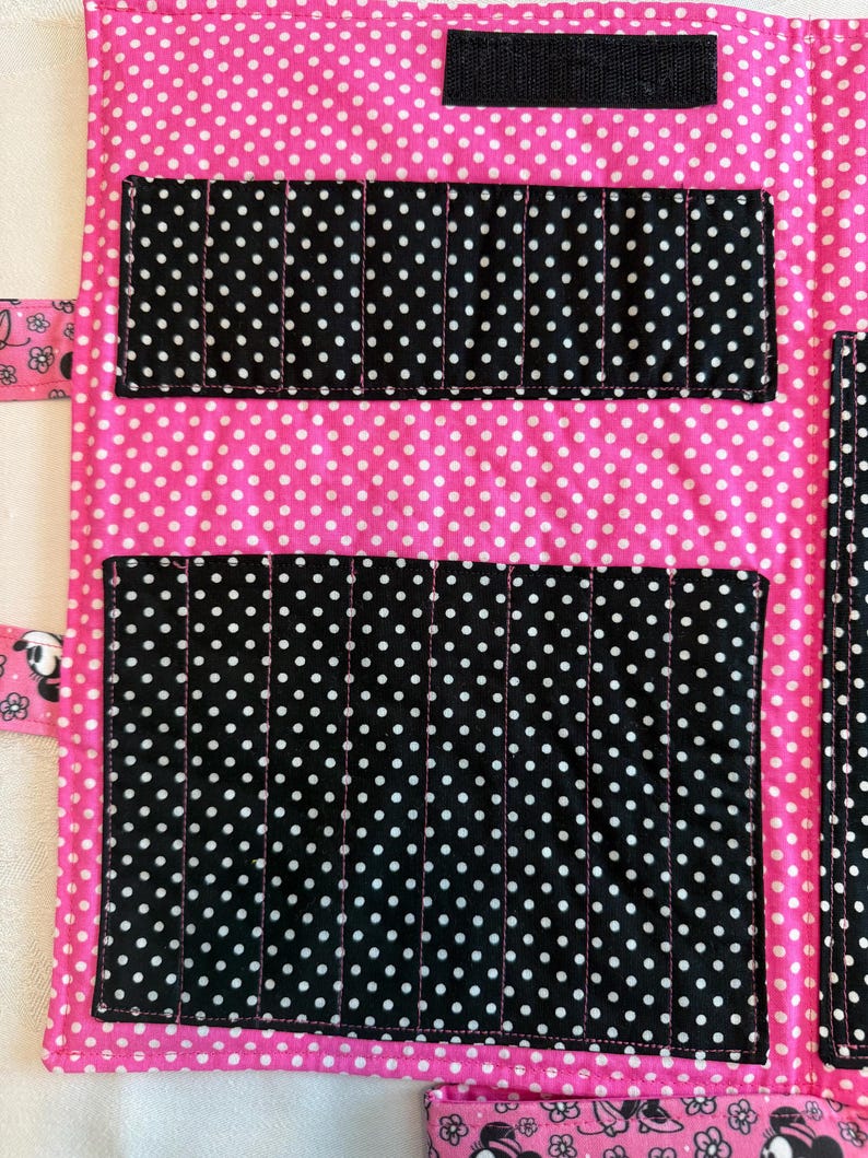Pu&ograve; includere: Un organizer in tessuto con una base rosa brillante a pois bianchi. Include diverse tasche nere a pois bianchi per riporre oggetti, una striscia di velcro nera nella parte superiore e un bordo rosa con un motivo floreale in bianco e nero.