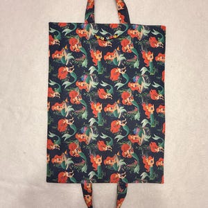 May include: A rectangular fabric tote bag featuring a dark blue background with a repeating pattern of the mermaid character Ariel with red hair and a green tail. The bag includes two fabric handles and a small string of letter beads spelling ELVAL.