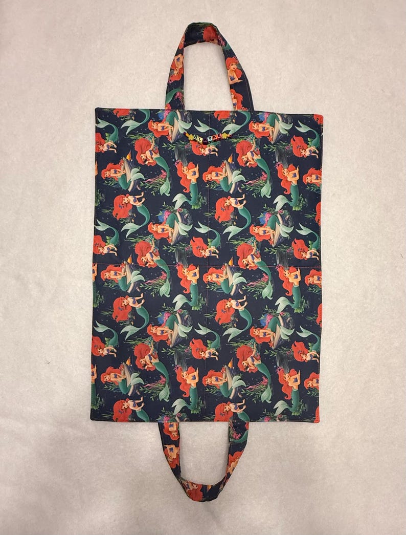 May include: A rectangular fabric tote bag featuring a dark blue background with a repeating pattern of the mermaid character Ariel with red hair and a green tail. The bag includes two fabric handles and a small string of letter beads spelling ELVAL.