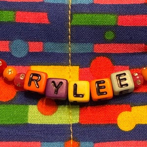 May include: A string of colorful plastic beads spelling the name RYLEE in black letters on square blocks. The beads are arranged in a horizontal line, featuring a mix of yellow, orange, red, purple, and white cubes, accented by smaller round beads.