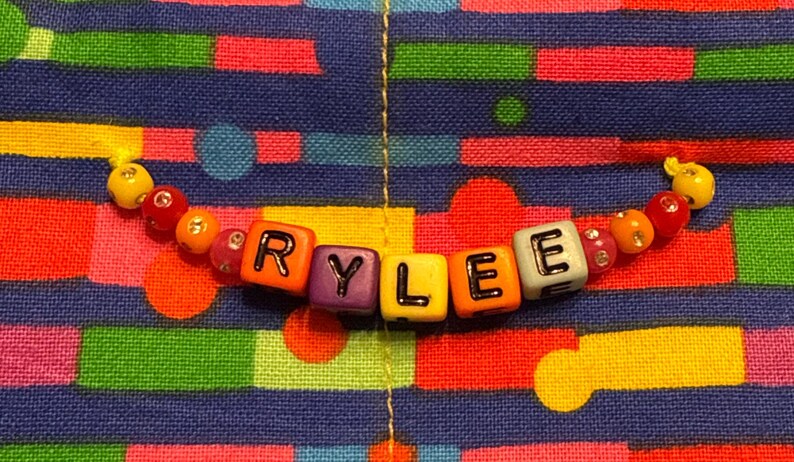 May include: A string of colorful plastic beads spelling the name RYLEE in black letters on square blocks. The beads are arranged in a horizontal line, featuring a mix of yellow, orange, red, purple, and white cubes, accented by smaller round beads.