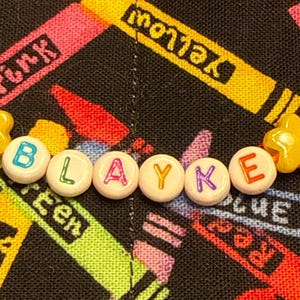 May include: A beaded bracelet featuring white round beads with colorful letters spelling the name BLAYKE, flanked by two yellow star-shaped beads on an orange string. The beads are arranged in a horizontal line against a black fabric background.