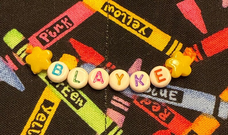 May include: A beaded bracelet featuring white round beads with colorful letters spelling the name BLAYKE, flanked by two yellow star-shaped beads on an orange string. The beads are arranged in a horizontal line against a black fabric background.