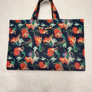 May include: A rectangular fabric tote bag featuring a dark blue background with a repeating pattern of a red-haired mermaid. The bag has two top handles and a small string of colorful letter beads spelling SLAYKE attached to the front.
