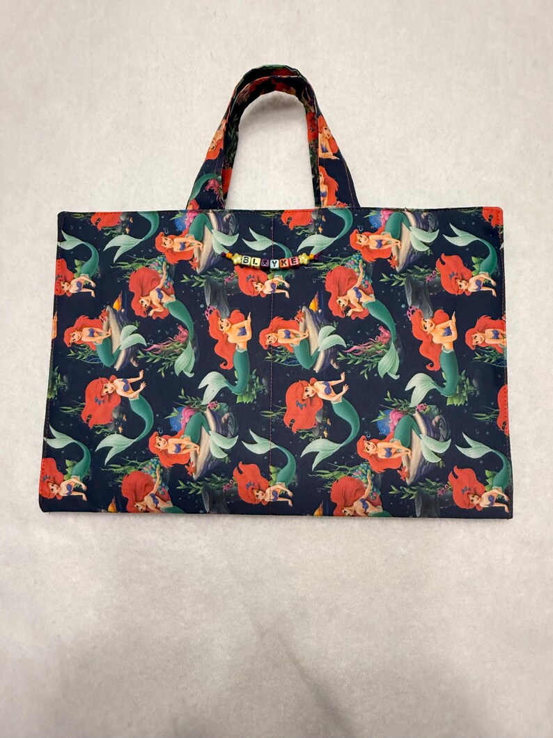 May include: A rectangular fabric tote bag featuring a dark blue background with a repeating pattern of a red-haired mermaid. The bag has two top handles and a small string of colorful letter beads spelling SLAYKE attached to the front.