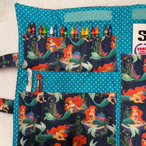 May include: A fabric crayon roll organizer featuring a teal background with white polka dots and a dark blue pocket fabric printed with Ariel the mermaid. The organizer contains multiple crayons, two pens, and a white card with the text BONUS STAND.