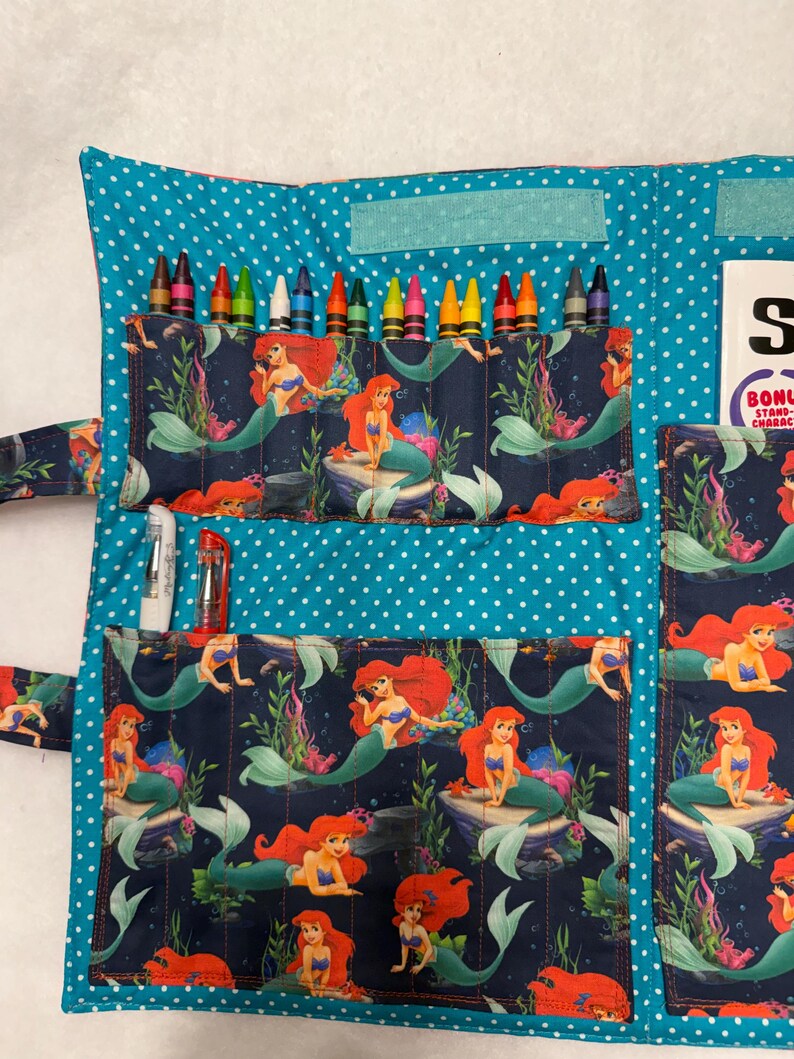 May include: A fabric crayon roll organizer featuring a teal background with white polka dots and a dark blue pocket fabric printed with Ariel the mermaid. The organizer contains multiple crayons, two pens, and a white card with the text BONUS STAND.