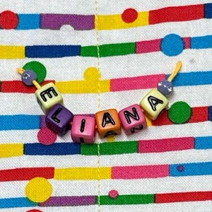 May include: A string of colorful plastic cube beads spelling the name ELIANA in black letters. The beads are yellow, purple, pink, orange, and pink, with small purple spherical beads at each end, all threaded on a thin yellow cord.