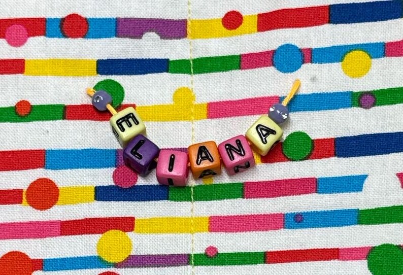 May include: A string of colorful plastic cube beads spelling the name ELIANA in black letters. The beads are yellow, purple, pink, orange, and pink, with small purple spherical beads at each end, all threaded on a thin yellow cord.