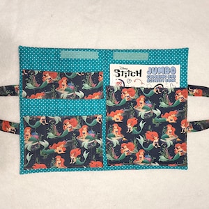 May include: A teal fabric organizer with white polka dots featuring three pockets and two side handles. The pockets are decorated with a dark blue pattern of red-haired mermaids. A Disney Stitch Jumbo Coloring and Activity Book is tucked inside.