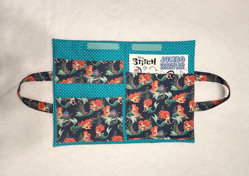 May include: A teal fabric organizer with white polka dots featuring three pockets and two side handles. The pockets are decorated with a dark blue pattern of red-haired mermaids. A Disney Stitch Jumbo Coloring and Activity Book is tucked inside.