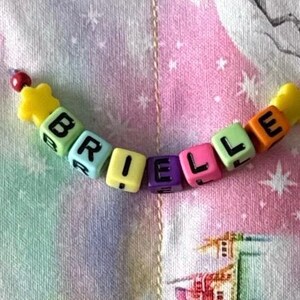 May include: A string of colorful plastic cube beads spelling the name BRIELLE in black capital letters. The beads are arranged in a curved line, featuring yellow, light green, light blue, purple, pink, and orange colors, with yellow star-shaped beads at both ends.