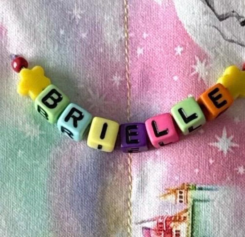 May include: A string of colorful plastic cube beads spelling the name BRIELLE in black capital letters. The beads are arranged in a curved line, featuring yellow, light green, light blue, purple, pink, and orange colors, with yellow star-shaped beads at both ends.