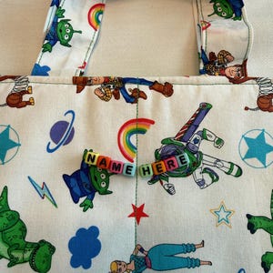 May include: A white fabric tote bag featuring a repeating pattern of cartoon characters including a green alien, a space ranger, a cowboy, and a shepherdess, along with rainbows, stars, and planets. The text NAME HERE is spelled out in colorful beads.