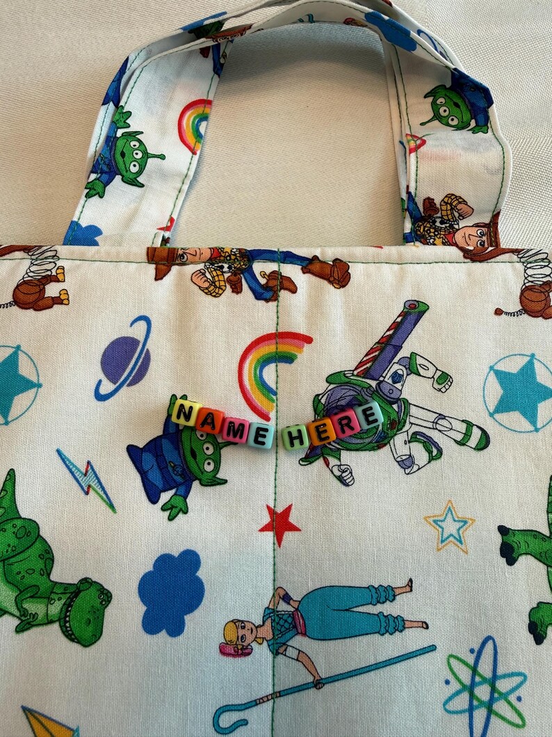 May include: A white fabric tote bag featuring a repeating pattern of cartoon characters including a green alien, a space ranger, a cowboy, and a shepherdess, along with rainbows, stars, and planets. The text NAME HERE is spelled out in colorful beads.