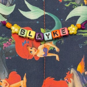 May include: A beaded name bracelet featuring the name BLAYKE spelled out in colorful square plastic letter beads, accented with yellow star-shaped beads and orange round beads, set against a dark blue fabric with a mermaid print pattern.