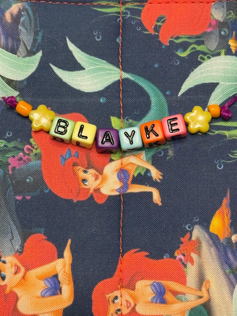May include: A beaded name bracelet featuring the name BLAYKE spelled out in colorful square plastic letter beads, accented with yellow star-shaped beads and orange round beads, set against a dark blue fabric with a mermaid print pattern.