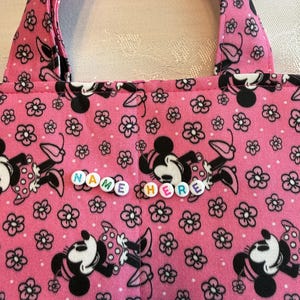 Pu&ograve; includere: Una borsa in tessuto rosa con un motivo ripetuto di topi dei cartoni animati in bianco e nero e piccoli fiori neri. La borsa include due manici in tessuto e una fila orizzontale centrale di perline di plastica bianca.