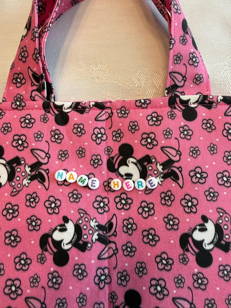 Pu&ograve; includere: Una borsa in tessuto rosa con un motivo ripetuto di topi dei cartoni animati in bianco e nero e piccoli fiori neri. La borsa include due manici in tessuto e una fila orizzontale centrale di perline di plastica bianca.