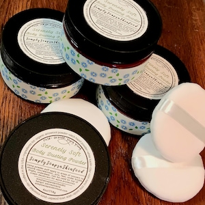 May include: Four round containers of Serenely Soft Body Dusting Powder with black lids and labels featuring text and floral patterns. Each container holds 4oz of powder. Two white circular powder puffs with satin ribbons are included in the set.