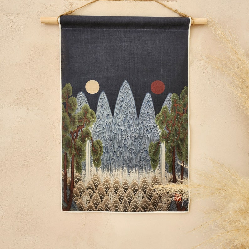 May include: A wall hanging featuring a landscape scene with mountains, trees, and two celestial bodies. The artwork is predominantly blue, green, and brown, with a dark blue background. The piece is mounted on a wooden rod and suspended by a natural fiber cord.