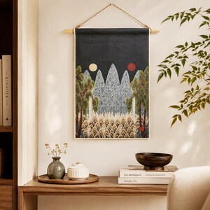 May include: A wall hanging with a dark blue background features a landscape design with mountains, trees, and two celestial bodies. The artwork is framed by a wooden rod and a natural fiber cord. The scene is complemented by a wooden table with decorative items.