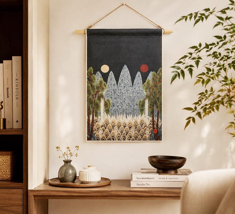 May include: A wall hanging with a dark blue background features a landscape design with mountains, trees, and two celestial bodies. The artwork is framed by a wooden rod and a natural fiber cord. The scene is complemented by a wooden table with decorative items.
