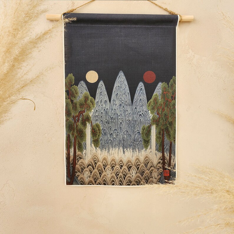 May include: A wall hanging featuring a landscape painting with mountains, trees, and two celestial bodies. The artwork is in shades of blue, green, and brown, with a dark background. The piece is suspended from a wooden rod with a jute cord.