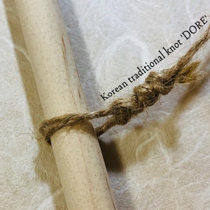 May include: Close-up of a wooden dowel tied with a natural fiber rope in a traditional Korean knot, labeled "DORE". The rope is wrapped around the dowel and knotted. The background is a textured, off-white surface.