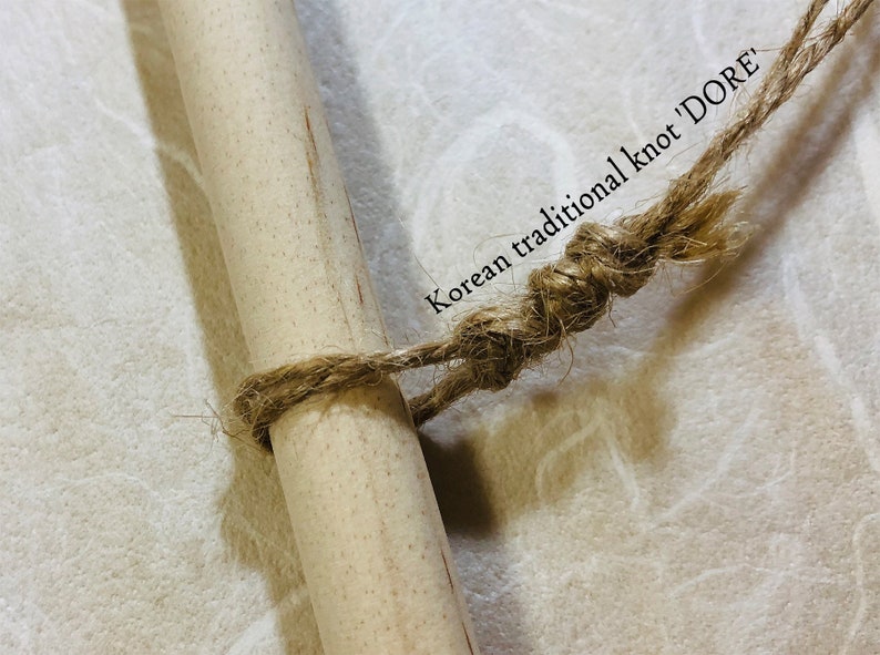 May include: Close-up of a wooden dowel tied with a natural fiber rope in a traditional Korean knot, labeled "DORE". The rope is wrapped around the dowel and knotted. The background is a textured, off-white surface.