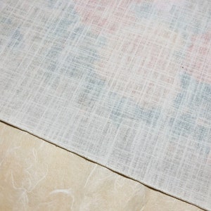 May include: Close-up of a white linen fabric with a subtle crosshatch pattern. Hints of red and blue floral designs are visible beneath the surface. The fabric is folded over a light beige surface.